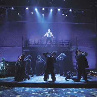 An a scene from ɫ's presentation of the musical Spring Awakening, an ensemble of actors in period costume kneel while a male soloist sings under a blue spotlight on an elevated platform.