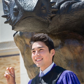 A male ɫ student in Honors regalia makes the two-fingered "Go Frogs" hand sign in front of a bronze statue