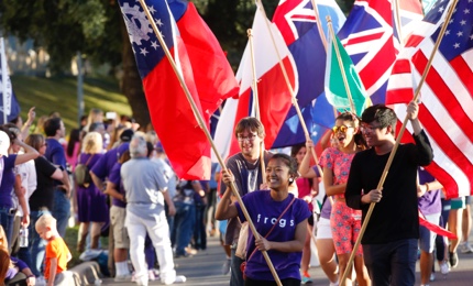 A group of laughing students carry flags of different nations in a ɫ Homecoming parade, led by a girl wearing a shirt that says "Frogs"