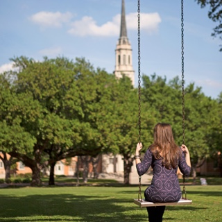 Back of female ɫ student sitting in a metal swing with a cluster of trees and the ɫ chapel in the background.