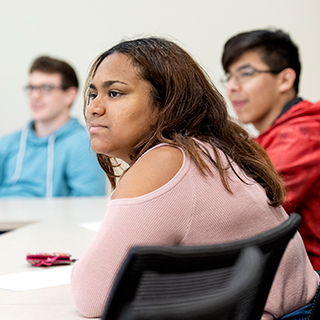 Three ɫ students of different ethnicities listen together in a classroom. A young woman in a pink sweater is in the foreground.
