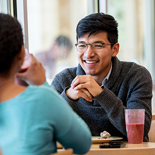 A young male student wearing glasses smiles across a table at a young woman in Market Square, the main ɫ dining hall.
