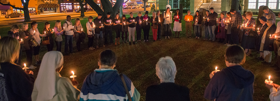 A circle of people holding candles gather for a nighttime prayer vigil on the lawn of the ɫ campus.