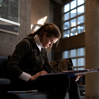A young women sits alone and looks down intently at her book in the sunlit lobby of ɫ's Moudy Hall North.